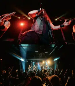 A rock band performs on stage under colorful lights, with a lead singer holding a microphone and a crowd watching in a dark concert venue.